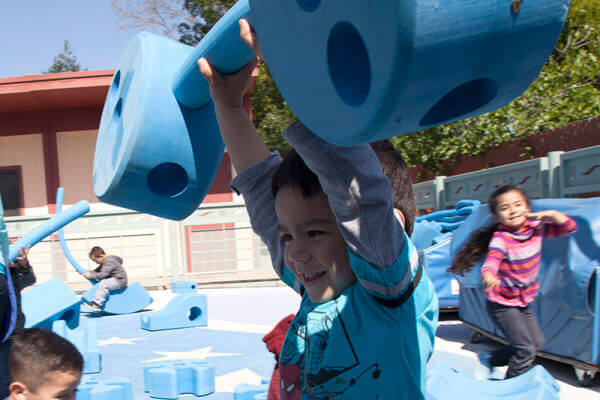 A smiling child lifting blue playground equipment overhead, similar to lifting weights